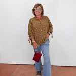 A woman stands against a white wall wearing a brown and gold patterned button-up blouse with tassel ties, light blue flare jeans, and dark sandals, while holding a small burgundy handbag.