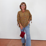 A woman stands against a white wall wearing a brown and gold patterned button-up blouse with tassel ties, light blue flare jeans, and dark sandals, while holding a small burgundy handbag.