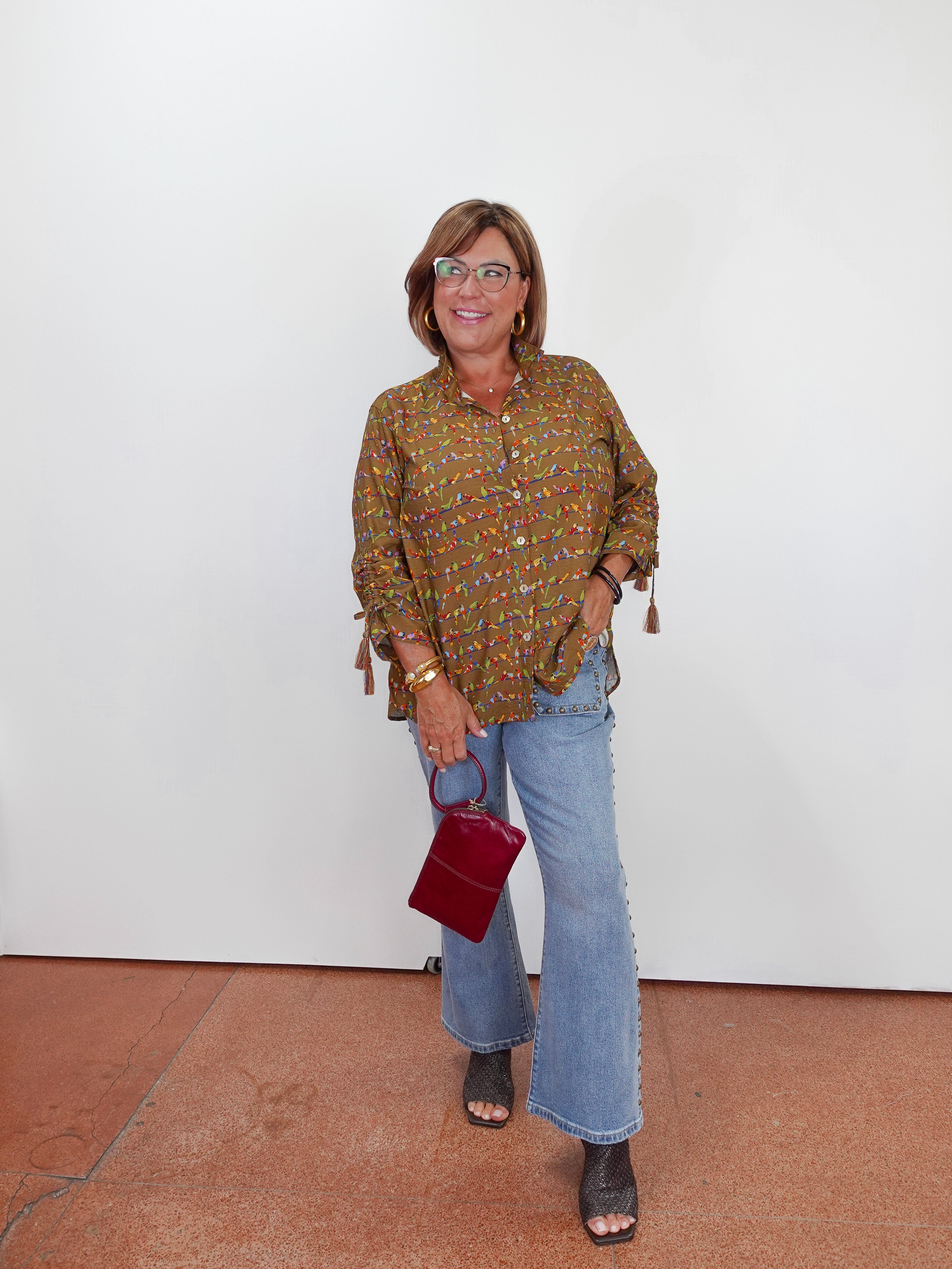 A woman stands against a white wall wearing a brown and gold patterned button-up blouse with tassel ties, light blue flare jeans, and dark sandals, while holding a small burgundy handbag.