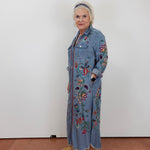 An older woman with short white hair stands against a plain white wall wearing a long-sleeve, button-front denim-style maxi dress with colorful floral embroidery and tan slide sandals.