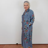 An older woman with short white hair stands against a plain white wall wearing a long-sleeve, button-front denim-style maxi dress with colorful floral embroidery and tan slide sandals.