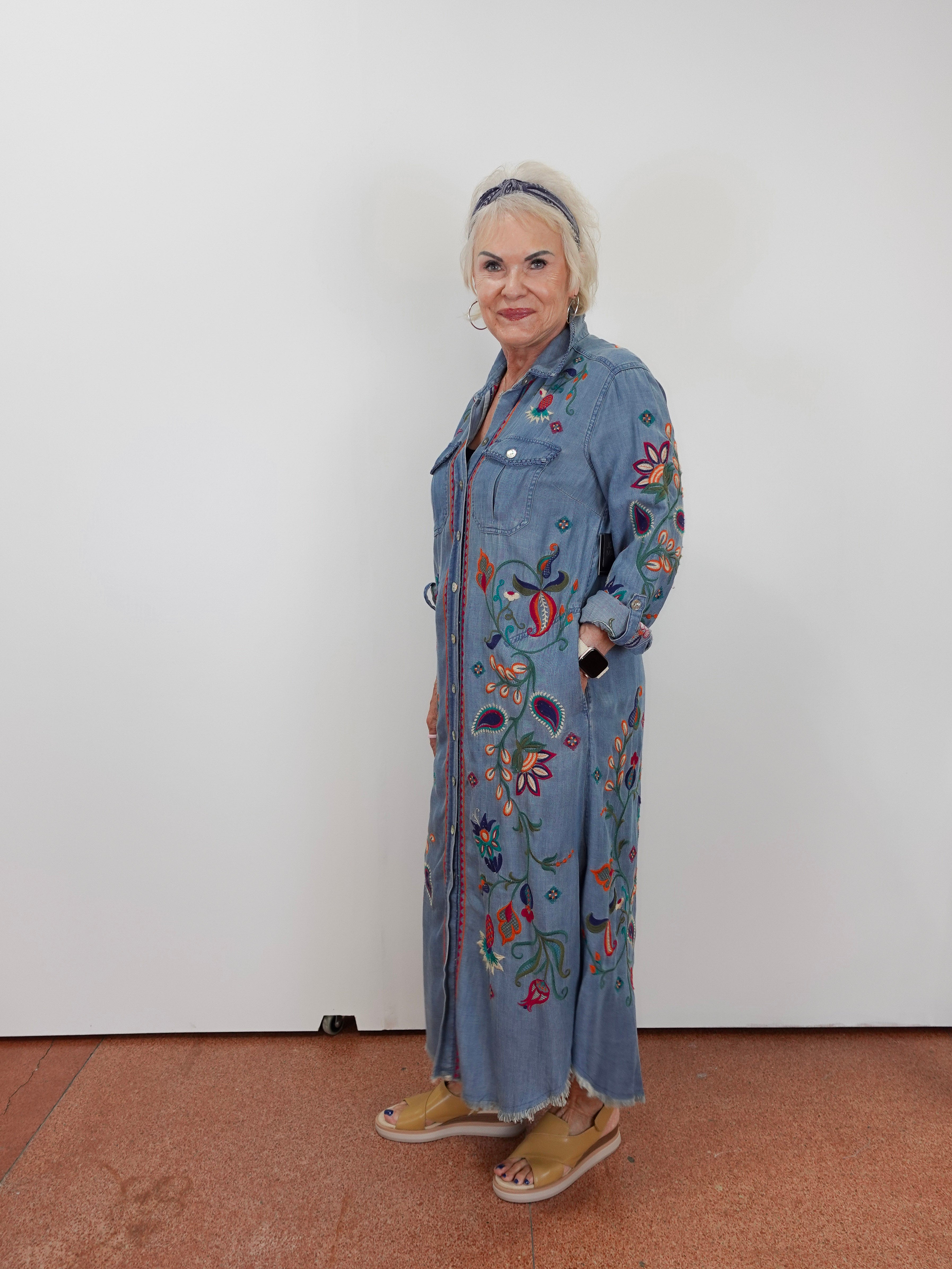 An older woman with short white hair stands against a plain white wall wearing a long-sleeve, button-front denim-style maxi dress with colorful floral embroidery and tan slide sandals.