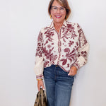 Woman wearing a floral blouse and jeans, holding a handbag against a white wall.