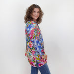 Woman wearing a colorful floral blouse and blue jeans against a white wall.