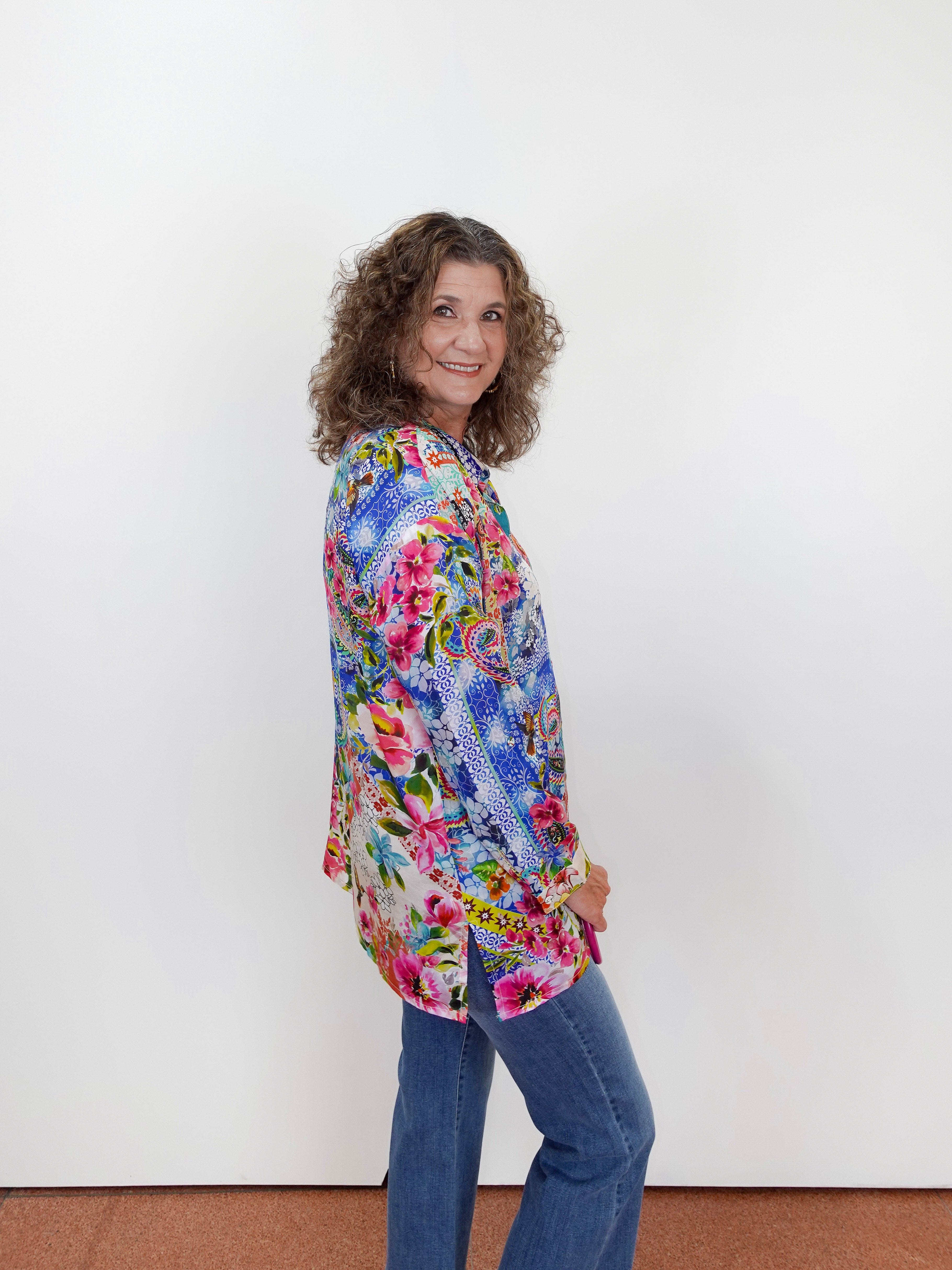 Woman wearing a colorful floral blouse and blue jeans against a white wall.