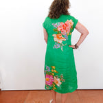 Woman wearing a green dress with floral patterns standing against a white wall.