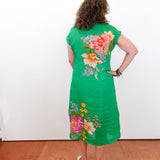 Woman wearing a green dress with floral patterns standing against a white wall.
