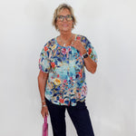Woman wearing a colorful floral blouse and holding a pink bag against a white wall.