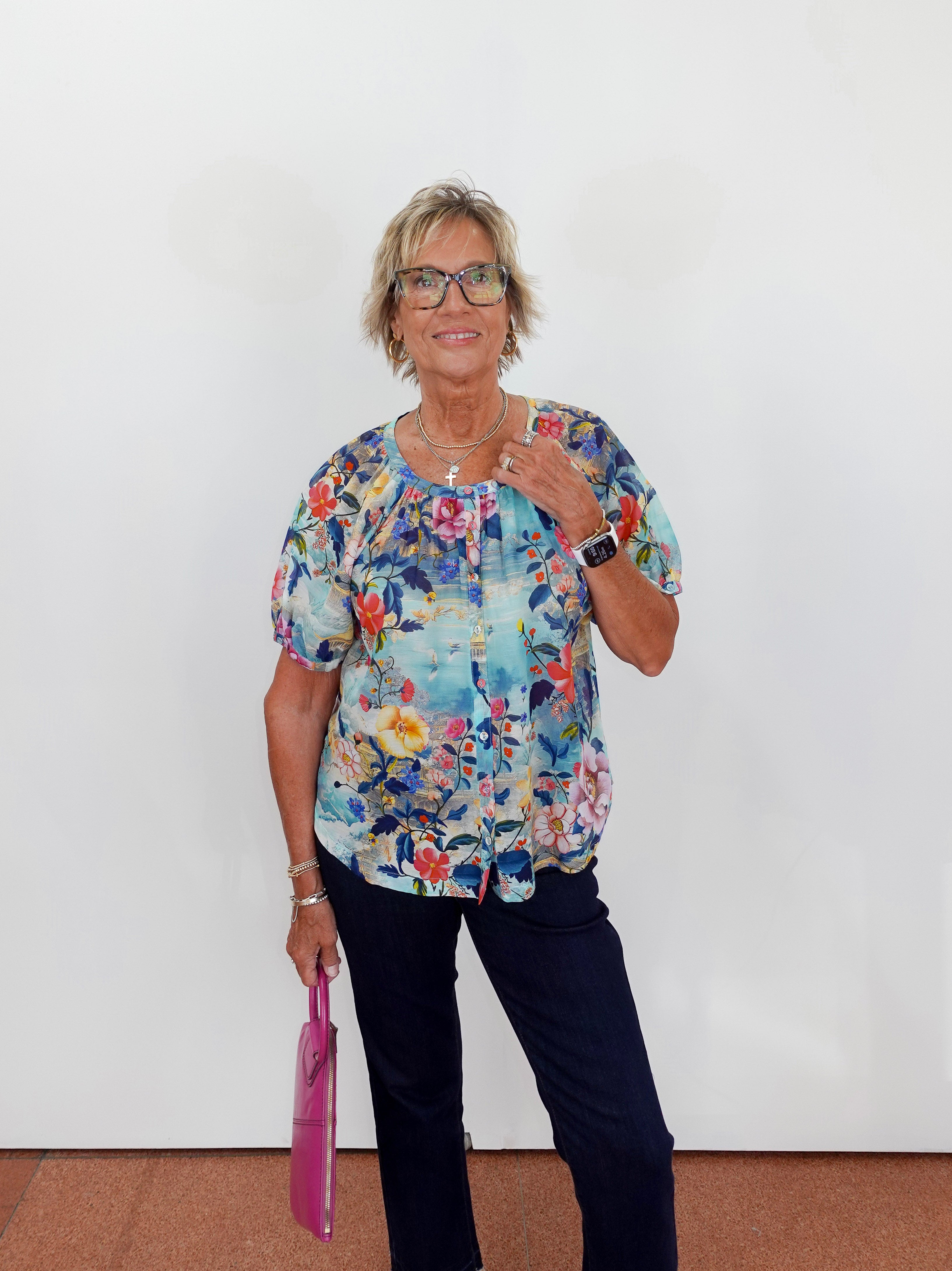 Woman wearing a colorful floral blouse and holding a pink bag against a white wall.