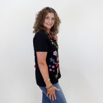 Woman wearing a black t-shirt with colorful floral designs and blue jeans against a white wall.