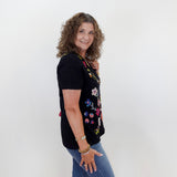 Woman wearing a black t-shirt with colorful floral designs and blue jeans against a white wall.