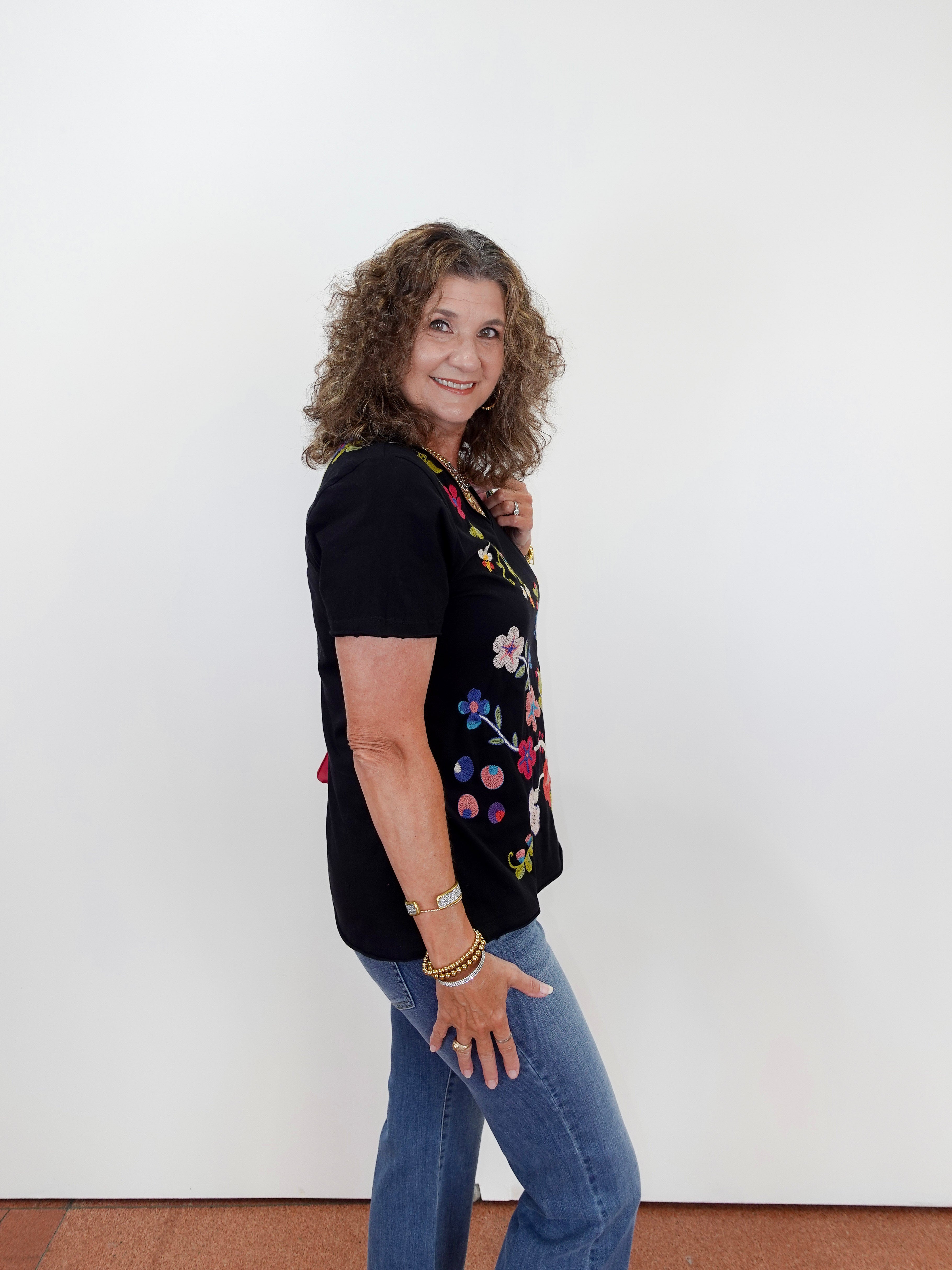 Woman wearing a black t-shirt with colorful floral designs and blue jeans against a white wall.
