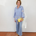Woman wearing a blue shirt with embroidered designs and jeans, holding a yellow clutch against a white wall.