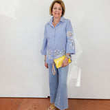 Woman wearing a blue shirt with embroidered designs and jeans, holding a yellow clutch against a white wall.