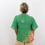 Person wearing a green blouse with floral designs on the back, standing against a white background