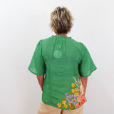 Person wearing a green blouse with floral designs on the back, standing against a white background