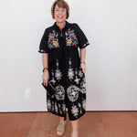 Woman wearing a black dress with floral patterns against a white wall.