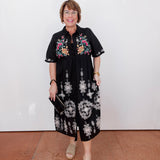 Woman wearing a black dress with floral patterns against a white wall.