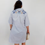 Person wearing a light blue dress with floral embroidery on the back, standing against a white wall.