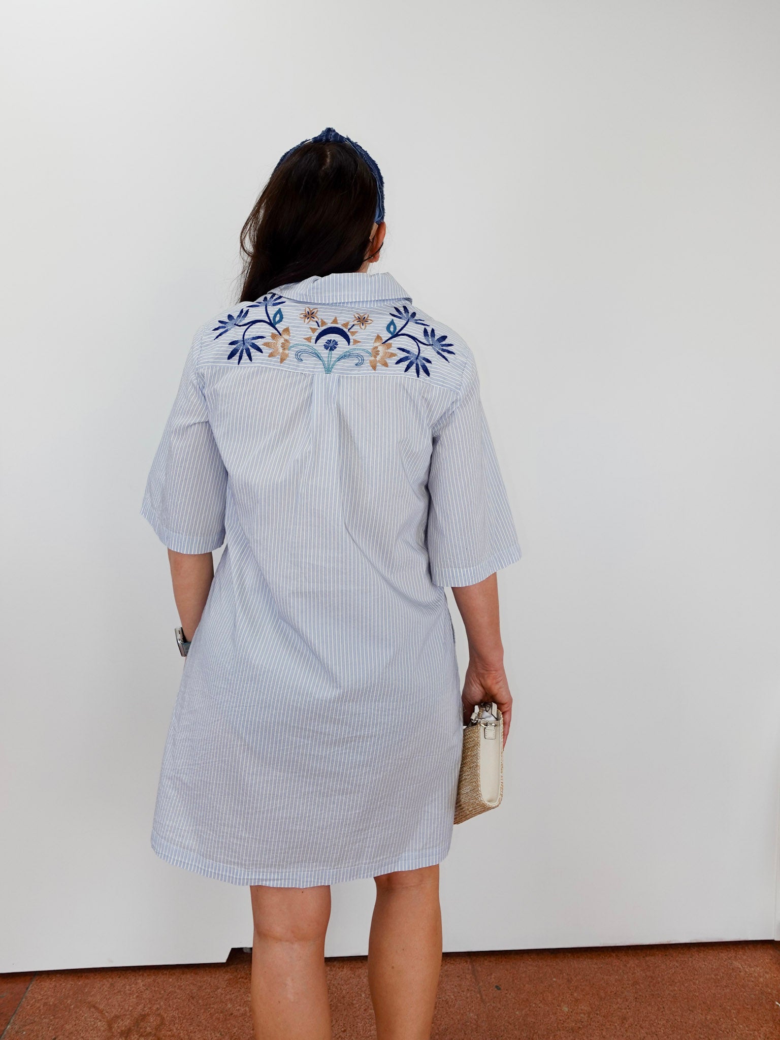 Person wearing a light blue dress with floral embroidery on the back, standing against a white wall.