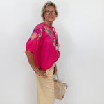 Woman wearing a pink embroidered blouse and beige pants against a white background