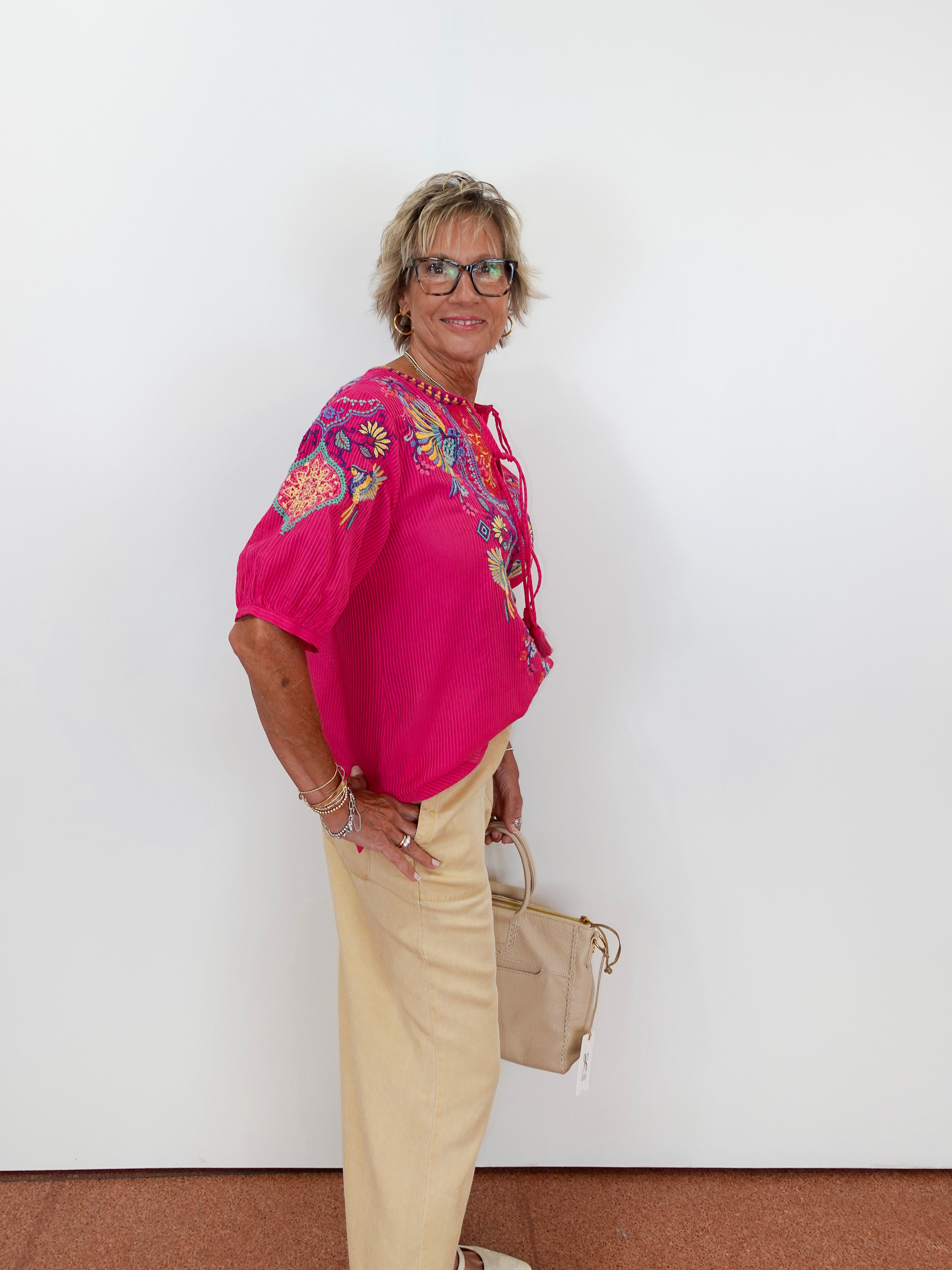 Woman wearing a pink embroidered blouse and beige pants against a white background
