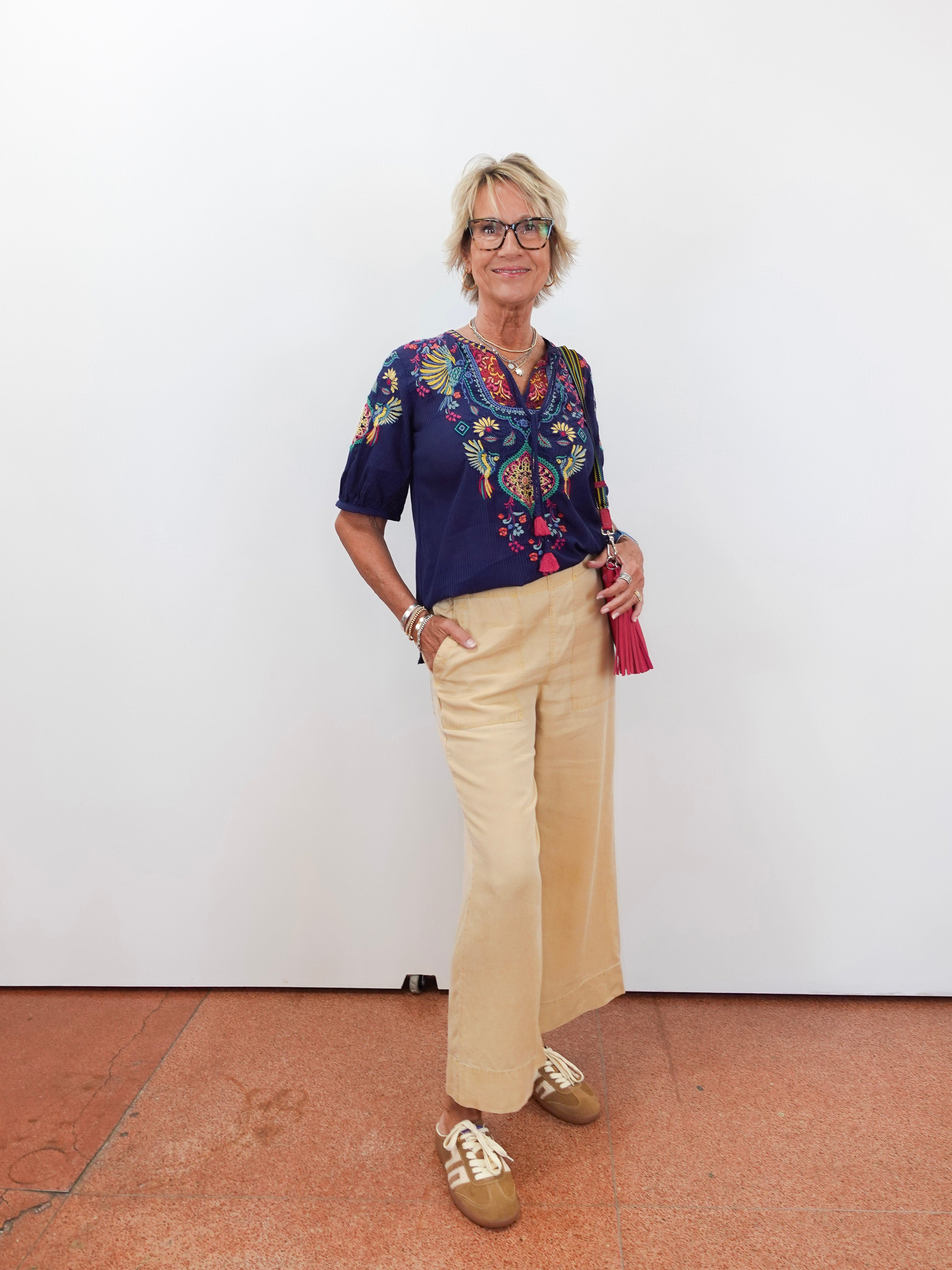 Person wearing a colorful blouse and beige pants against a white wall.