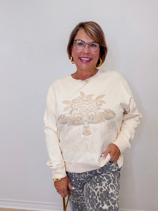 Woman wearing a cream sweatshirt with decorative elements against a white wall