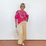 Woman wearing a bright pink blouse with floral patterns and beige pants, standing against a white wall.