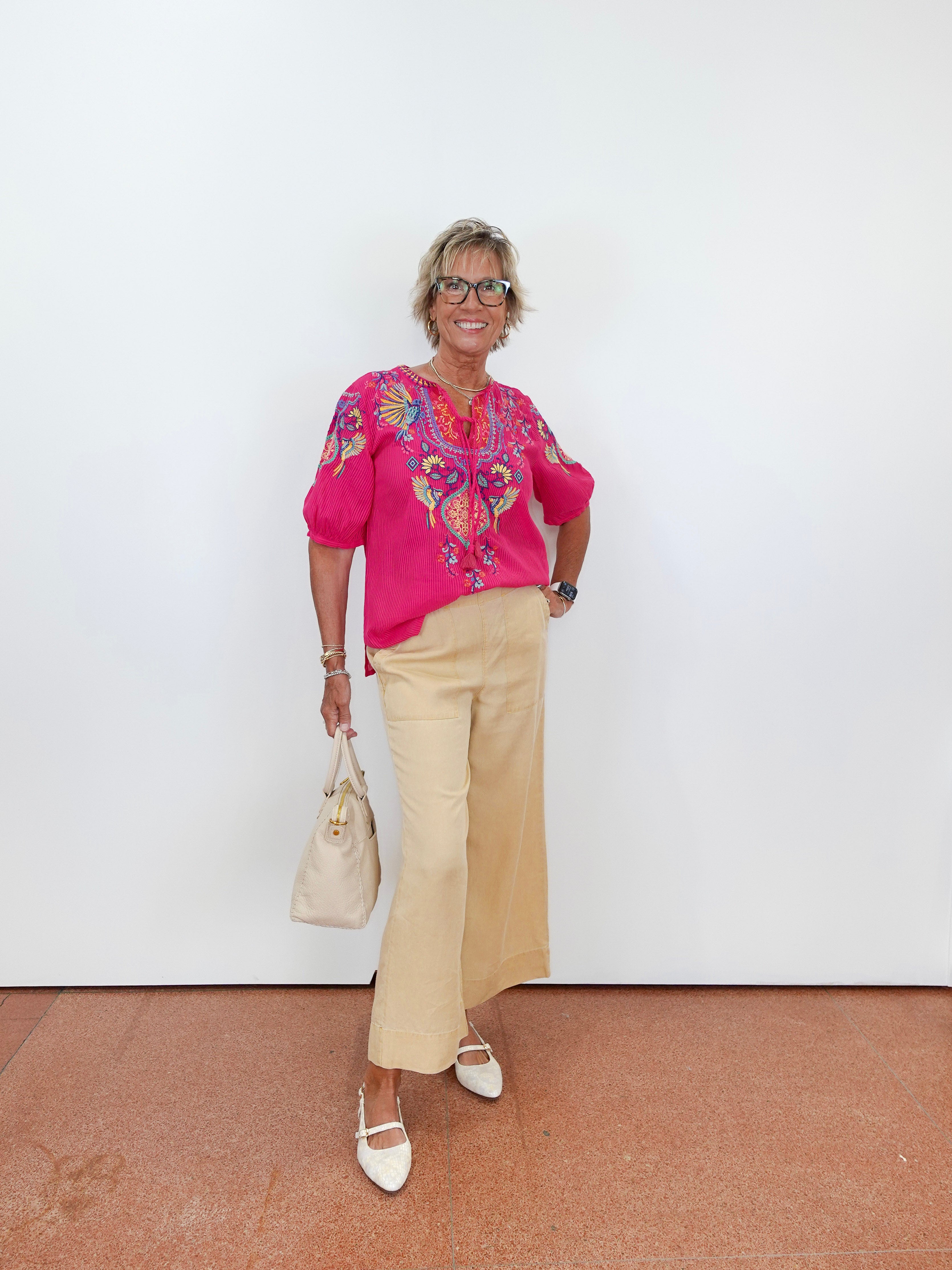 Woman wearing a bright pink blouse with floral patterns and beige pants, standing against a white wall.