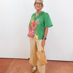 Woman wearing a green floral blouse with beige pants and holding a brown bag against a white wall.