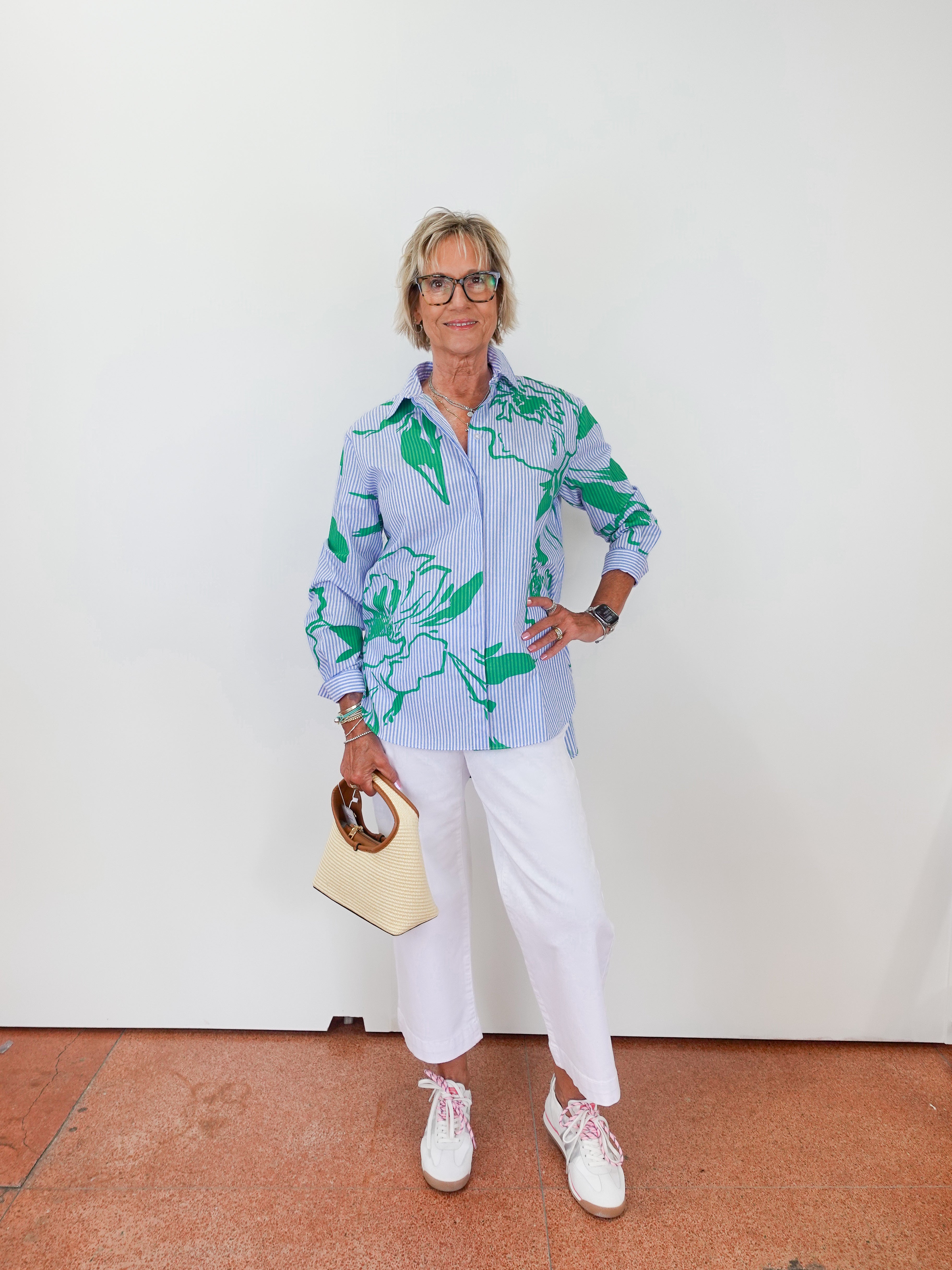 Woman wearing a light blue floral shirt with white pants, holding a beige handbag.