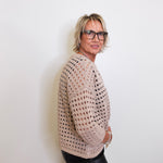 Woman wearing a beige open-knit sweater and black pants against a white background