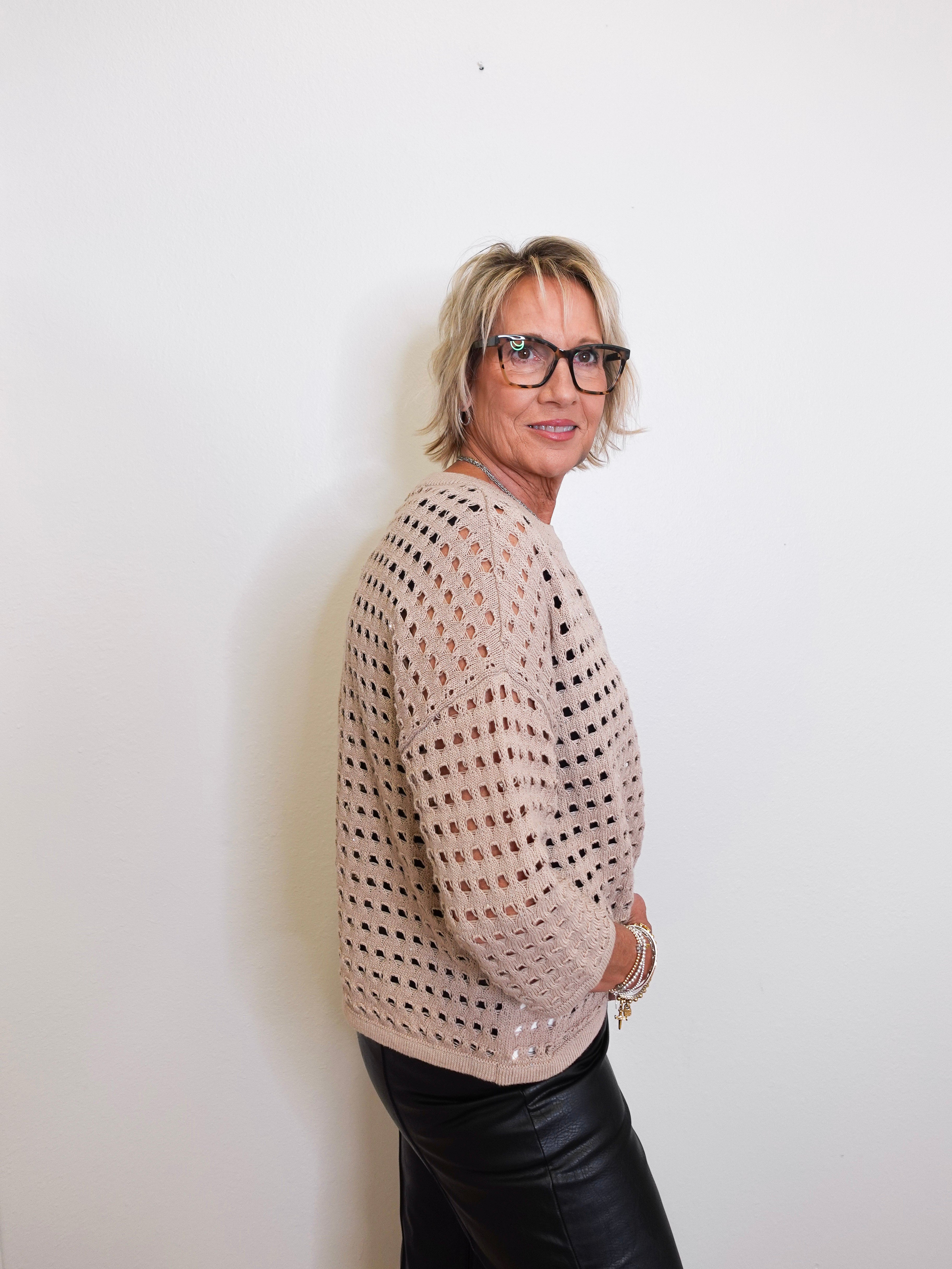 Woman wearing a beige open-knit sweater and black pants against a white background