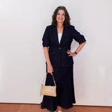 Woman in a navy suit with a beige handbag standing against a white wall.