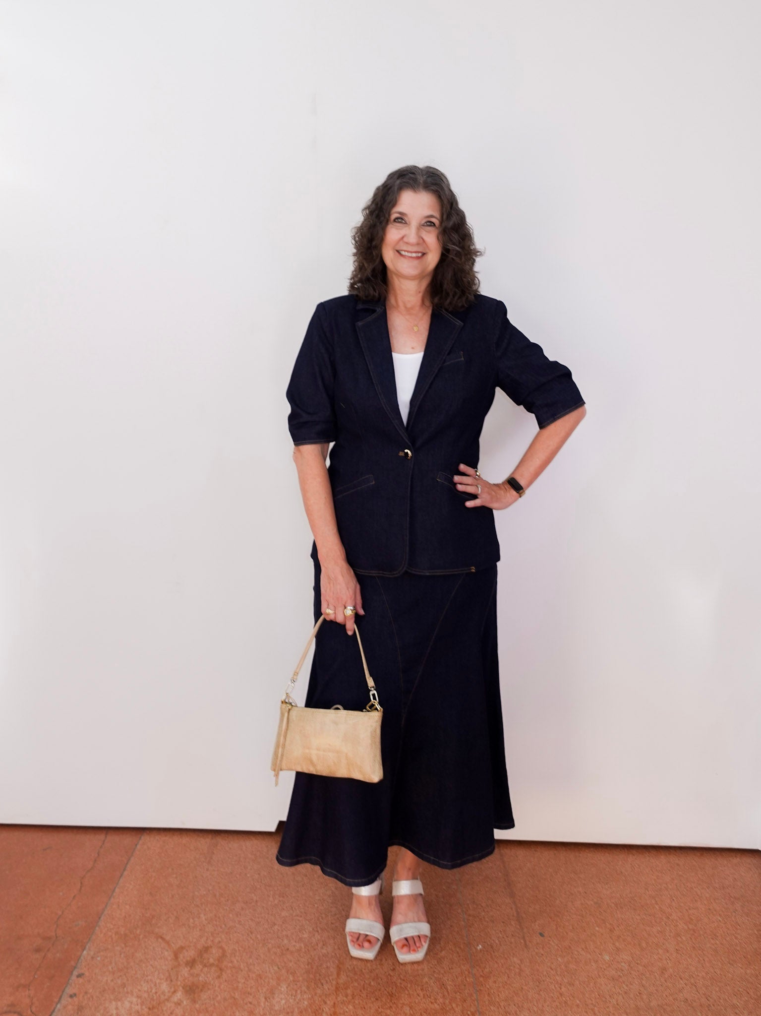 Woman in a navy suit with a beige handbag standing against a white wall.