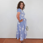 Woman wearing a blue floral dress standing against a white wall.