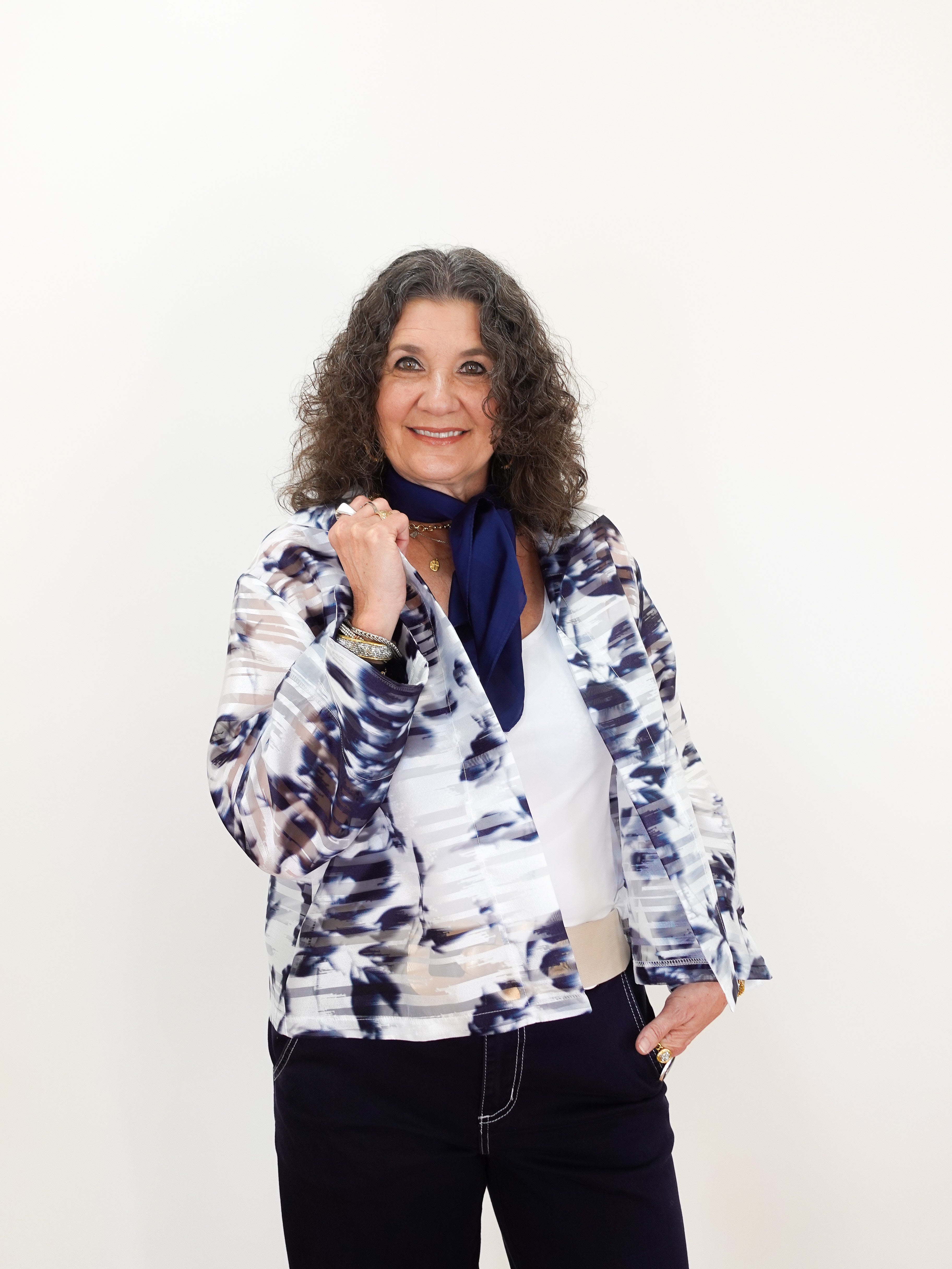 Woman wearing a patterned jacket over a white shirt with a blue scarf, standing against a plain background.