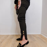 Person wearing black checkered pants with gold buttons and black loafers on a light wooden floor.