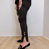 Person wearing black checkered pants with gold buttons and black loafers on a light wooden floor.