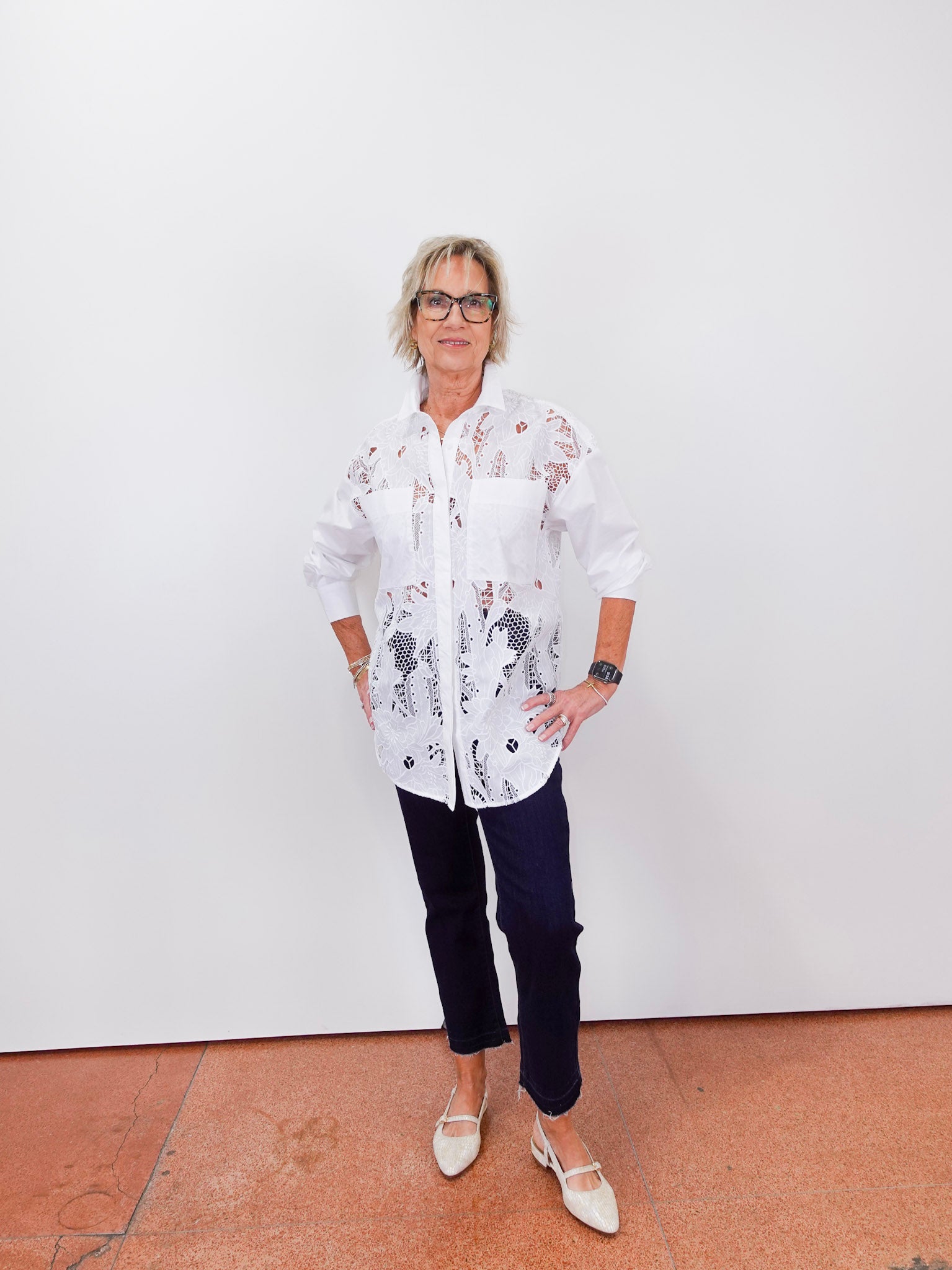 Person wearing a white floral shirt and dark pants against a white wall.