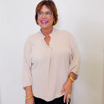 Woman wearing a beige blouse and black pants against a white wall