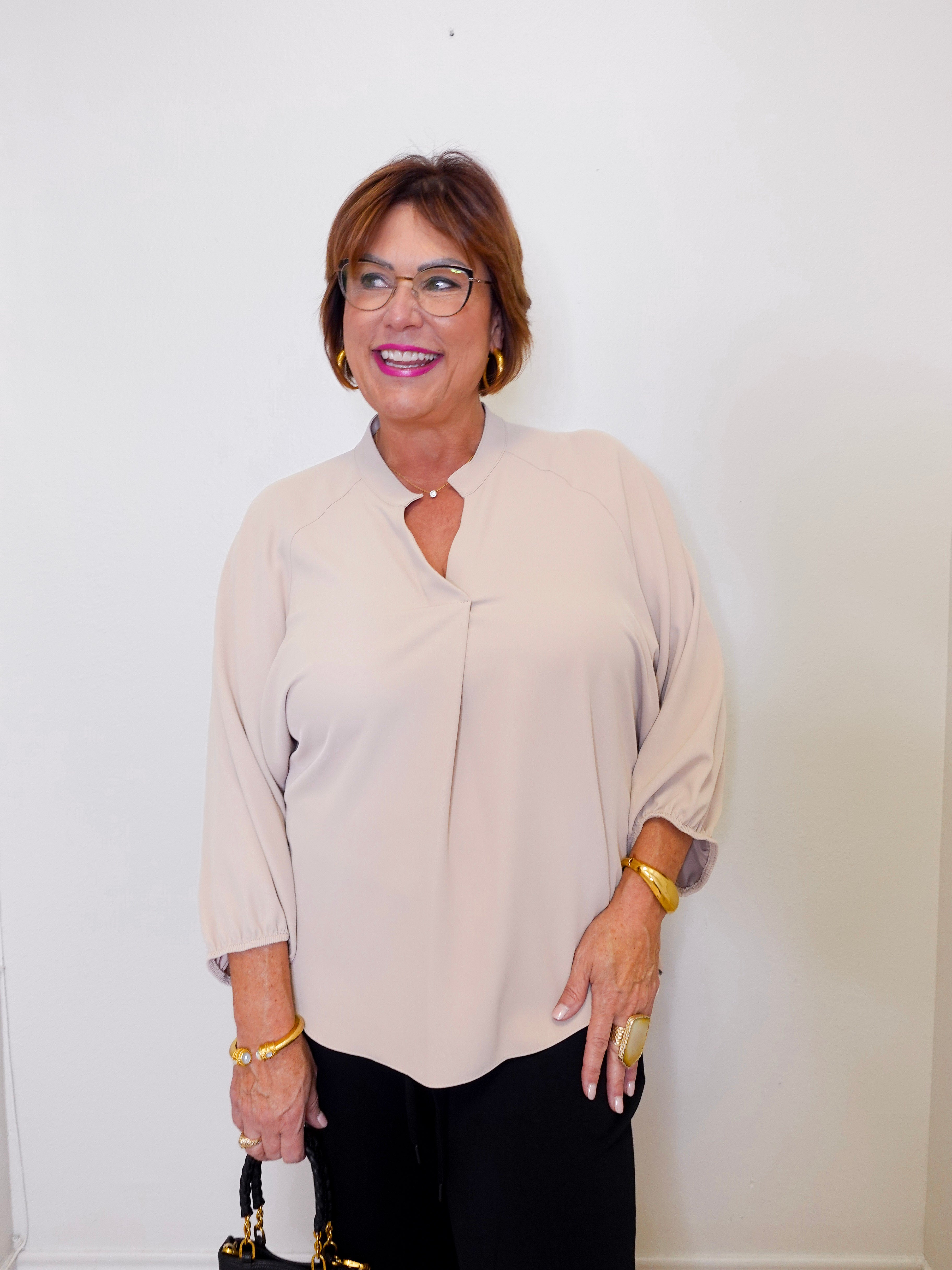 Woman wearing a beige blouse and black pants against a white wall