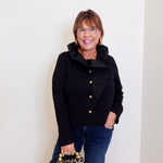 Woman wearing a black jacket with gold buttons, holding a leopard print handbag against a white wall.