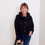 Woman wearing a black jacket with gold buttons, holding a leopard print handbag against a white wall.