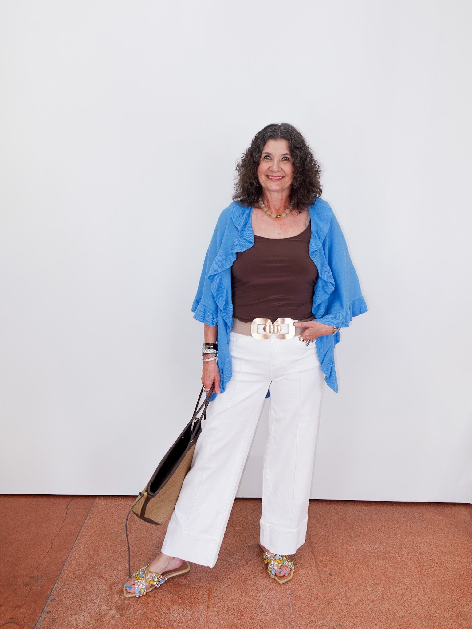 Woman wearing a blue jacket, brown top, white pants, and floral shoes against a white wall.