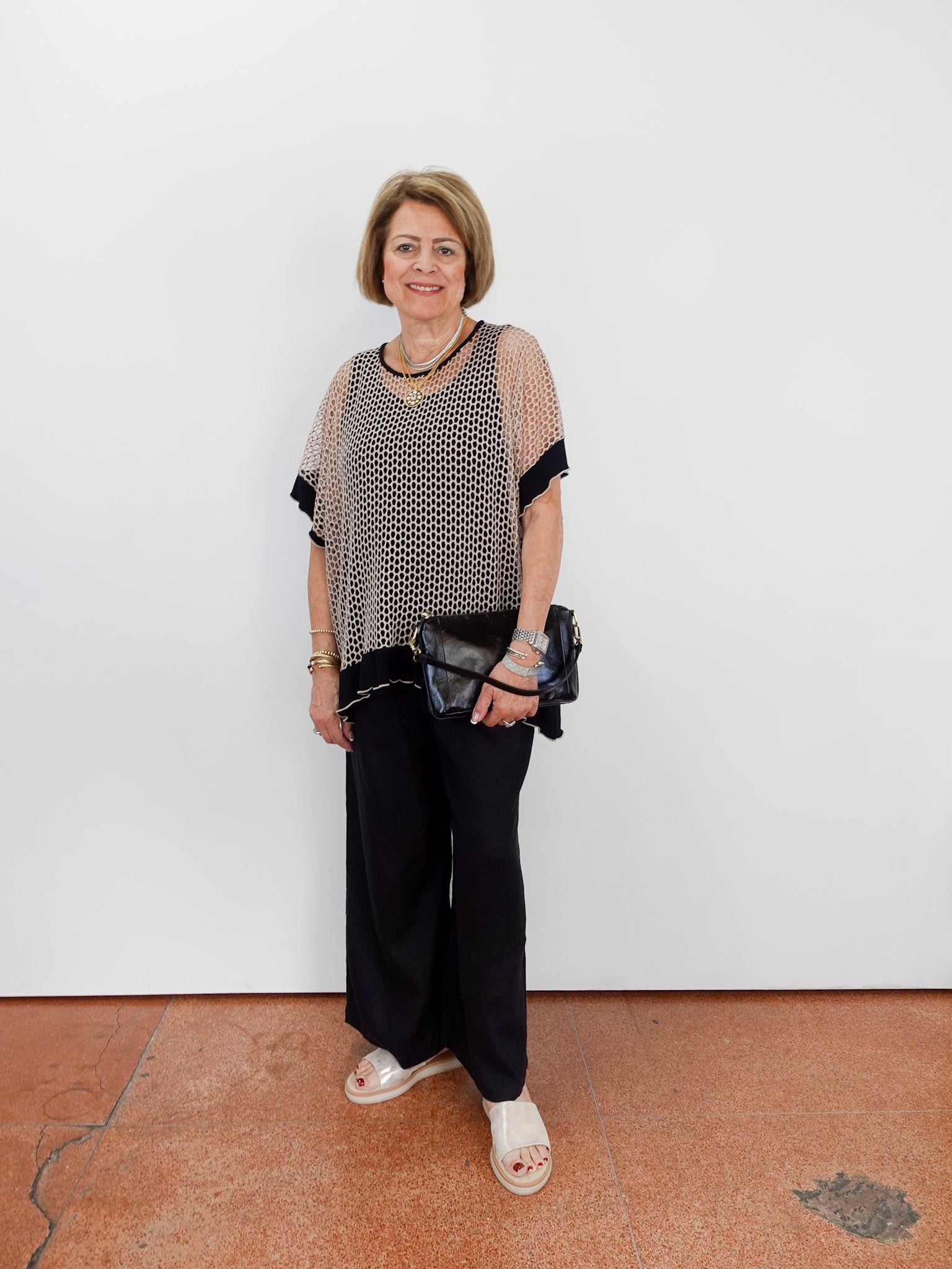 Woman wearing a patterned top and black pants against a white wall.