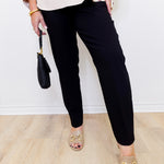 Person wearing black pants and gold sandals, holding a black handbag against a white background