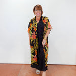 Woman wearing a black dress with a colorful floral kimono-style cover-up against a white wall.