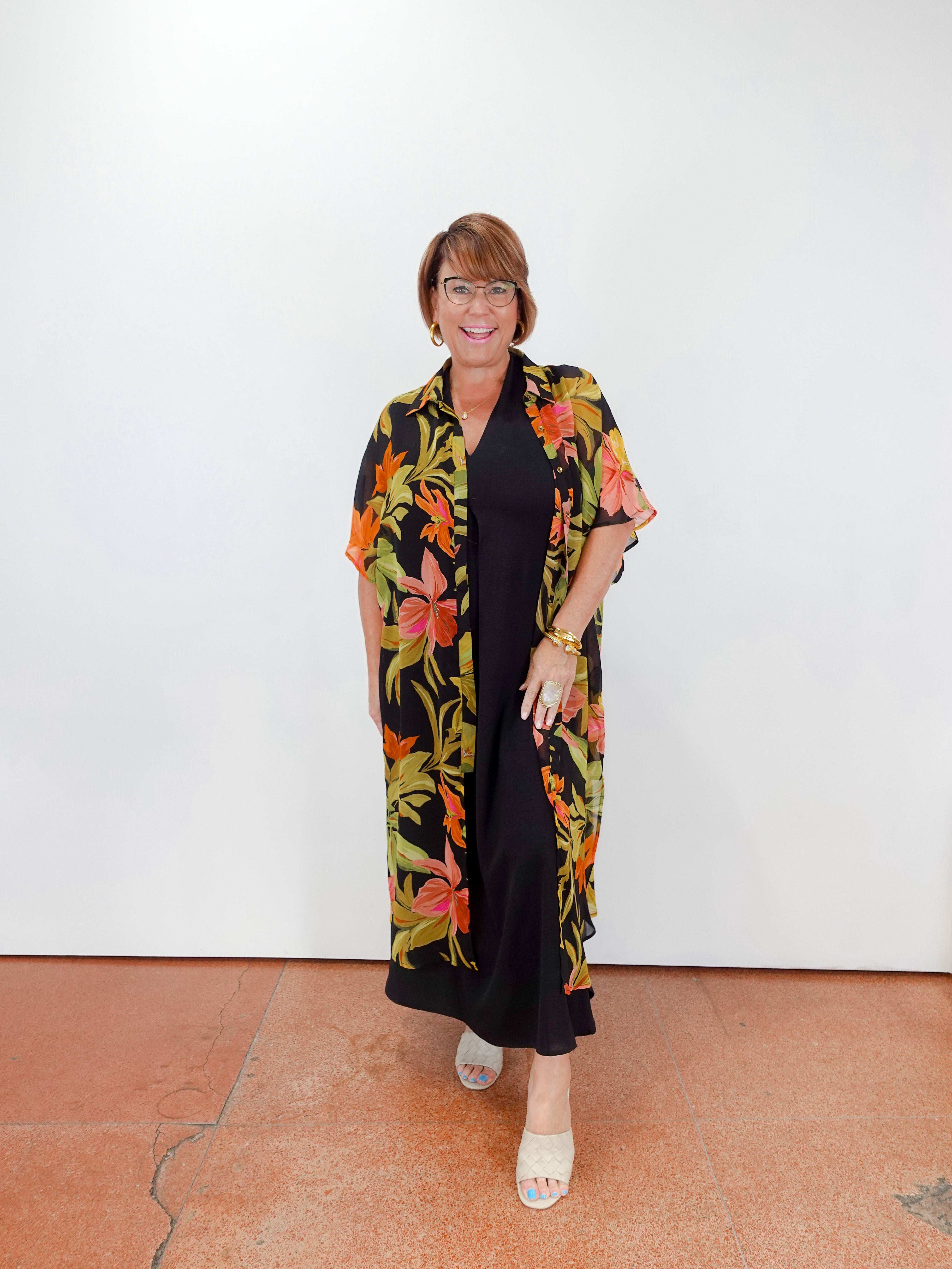 Woman wearing a black dress with a colorful floral kimono-style cover-up against a white wall.